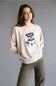 A woman wearing a light beige crewneck sweatshirt with a black and white dog graphic on the front, paired with olive green pants, against a solid gray studio background.
