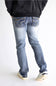 Rear view of a person wearing light-wash blue jeans with decorative embroidered detailing on the back pockets, paired with a black jacket and white sneakers, against a plain white studio background.
