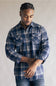 Model wearing the Split Men's long-sleeve flannel shirt in a navy blue, khaki, and white plaid pattern, featuring two chest flap pockets with button closures and buttoned cuffs against a grey studio background.