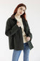 A woman with red hair wearing an olive green utility jacket with a brown corduroy collar, layered over a cream sweater and blue jeans, photographed against a plain white studio background.
