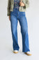 A person wearing blue wide-leg jeans with a faded wash, styled with an olive green jacket, a floral top, and white platform sneakers against a plain light gray studio background.