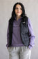 A woman wearing a black quilted vest over a purple quarter-zip sweatshirt and gray sweatpants, standing against a plain light gray background. The vest has a white The North Face logo on the chest.