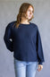 A woman wearing a dark navy blue waffle-knit long-sleeve sweater and light blue jeans, standing against a neutral gray studio background.