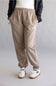 A person wearing beige jogger sweatpants with an elastic waistband and cuffs, styled with a white top, black jacket, and white sneakers with black stripes, shown from the waist down against a light gray studio background.