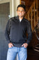 A smiling man wearing a dark charcoal gray quarter-zip pullover sweater with contrasting light gray shoulder panels and light blue jeans, standing in a home interior setting.