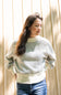 A woman wearing a cream-colored open-knit sweater with blue jeans, standing against a light wood-paneled wall.