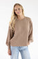 A woman with long, wavy blonde hair wearing a khaki crewneck ribbed knit top and light blue jeans against a white background.