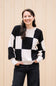 A woman wearing a black and off-white checkered knit sweater and dark jeans, standing against a beige corrugated metal wall.