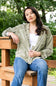 A woman sitting on a wooden bench outdoors, wearing an olive green corduroy button-up shirt over a white tank top and blue jeans, with a blurred green garden background.