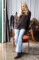 A woman stands in a rustic barn-like setting wearing a dark charcoal knit sweater, light blue flare jeans, and brown ankle boots, leaning against an orange chair.