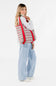 A woman modeling a large woven tote bag with a red, green, and white houndstooth pattern and solid red shoulder straps, paired with a white long-sleeve top, light blue wide-leg jeans, and white sneakers against a plain white studio background.