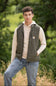 A young male model standing outdoors in a field, wearing the Carhartt Light Weight Insulated Vest for Men in Moss, layered over a cream hoodie sweatshirt and paired with light-wash blue jeans.