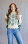 A smiling woman with long wavy red hair models a cream knit top with green floral embroidery and side ties, paired with blue jeans, against a plain gray studio background.