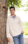 A young man smiling and leaning against a large tree trunk outdoors. He is wearing a light beige quarter-zip pullover sweater over a blue-gray t-shirt and blue jeans.