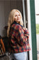 A woman with long blonde hair viewed from the back-side angle, wearing a burgundy, navy, and orange plaid barn jacket with a corduroy collar and light-wash denim jeans while looking back over her shoulder in a doorway.