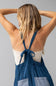 A woman viewed from behind wearing a blue polka-dot apron over a white ribbed tank top, with her arms raised, against a solid gray background.