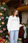 A woman posing in front of a decorated Christmas tree wearing the 1897 Active Merry and Bright Embroidered Fleece Sweatshirt in White with light-wash jeans.