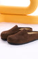 A pair of dark brown suede slip-on mule slippers with black soles, displayed on a white surface with a yellow abstract background prop.