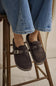 A person wearing blue jeans sits on a wooden stool, showing a pair of dark brown suede slip-on shoes with woven strap details and small silver buckle accents.