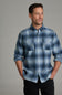A man wearing a blue and gray plaid button-down shirt with two chest pockets, paired with black pants, standing against a solid gray studio background.
