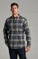 A man wearing a long-sleeve plaid button-up shirt in blue and tan, paired with dark jeans, standing against a solid gray studio background.