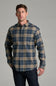 A man wearing a long-sleeve plaid button-up shirt in blue and tan, paired with dark jeans, standing against a solid gray studio background.