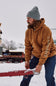 A man wearing a brown Carhartt hoodie and blue jeans stands in a snowy outdoor setting next to a horse-drawn sled, with the Carhartt brand name visible on the sleeve of the hoodie.