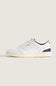 A low-top sneaker in white leather with navy blue accents on the collar, heel, and tongue, featuring a cream-colored rubber sole, shown against a solid light beige background.