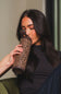 A woman with long dark hair holds a leopard-print insulated tumbler to her lips while wearing a black top in a softly lit indoor setting.
