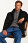A man wearing a black overshirt, gray henley shirt, and blue jeans sits on a chair against a plain white background.