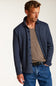 A man wearing a navy blue button-up knit overshirt over a taupe t-shirt and blue jeans, photographed against a clean off-white studio background.