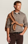 A man wearing a brown and beige checkered button-down shirt, dark brown trousers, and a brown knit sweater tied around his shoulders, photographed against a light gray studio background.