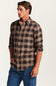 A man wearing a brown and beige buffalo plaid button-down flannel shirt with blue jeans, standing against a plain off-white background.