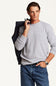 A man wearing a light gray long-sleeve knit crewneck sweater and blue jeans, holding a dark navy jacket over his shoulder against a plain off-white background.