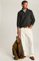 A man stands against a light gray studio background wearing a dark charcoal ribbed quarter-zip sweater over a light blue shirt, off-white trousers, and brown shoes. He holds a brown jacket in one hand.