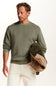 A man wearing an olive green crewneck sweater and off-white trousers, holding a dark green jacket with a beige sherpa lining against a plain white background.