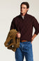 A man wearing a dark burgundy cable-knit quarter-zip sweater and blue jeans, holding a brown suede jacket against a plain light background.