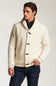 A man wearing a cream-colored textured knit cardigan with toggle closures and a shawl collar, paired with dark blue jeans and a dark plaid shirt, against a light gray studio background.