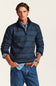 A man wearing a blue and navy patterned quarter-zip sweater over a light blue shirt and blue jeans, standing against a plain off-white background.
