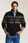 A man wearing a black zip-up knit sweater with a gray and white Fair Isle pattern across the chest and sleeves, layered over a light blue button-down shirt and blue jeans, against a plain light gray background.