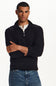 A man wearing a black ribbed quarter-zip mock neck sweater over a white collared shirt and blue jeans, standing with arms crossed against a plain white studio background.