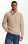 A man wearing a beige cable-knit sweater with a shawl collar, paired with blue jeans, photographed against a plain white studio background.