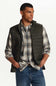A man wearing a dark olive green diamond-quilted zip-up vest over a gray and white plaid flannel shirt, paired with blue jeans, photographed against a clean white studio background.