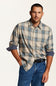A man wearing a beige and blue plaid button-down shirt with rolled-up sleeves, tucked into blue jeans with a black belt, against a plain white studio background.