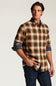 A man wearing a long-sleeve button-down plaid shirt in brown, orange, and green tones with rolled-up sleeves showing a blue undershirt, paired with dark blue jeans, against a light gray studio background.