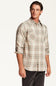 A man wearing a long-sleeve plaid button-up shirt in beige, brown, and white, paired with dark gray pants, photographed against a clean white studio background.