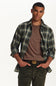 A man wearing a green, blue, and white plaid button-down shirt over a brown crewneck t-shirt, paired with dark green pants and a gold-tone belt, against a plain white background.