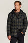A man wearing a dark navy and olive green quilted hooded jacket with a subtle plaid pattern, styled over a dark t-shirt and black trousers, photographed against a neutral light gray studio background.