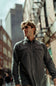 A man wearing a dark gray long-sleeve button-up shirt and sunglasses walking down an urban street.