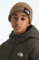 A person wearing a tan ribbed beanie with a black The North Face logo patch and an olive green puffer jacket with a white The North Face logo on the chest, photographed against a light gray studio background.